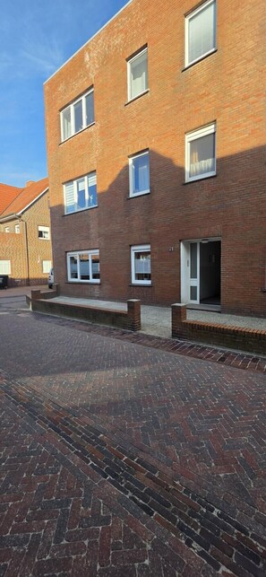 A multi-story brick building with multiple windows, a visible door, and a paved area in front.