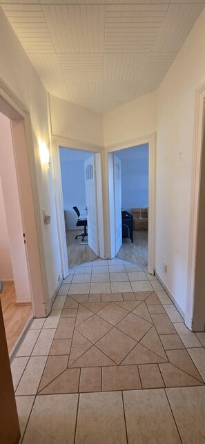 A hallway with a tiled floor, white walls, and a ceiling with wooden paneling.