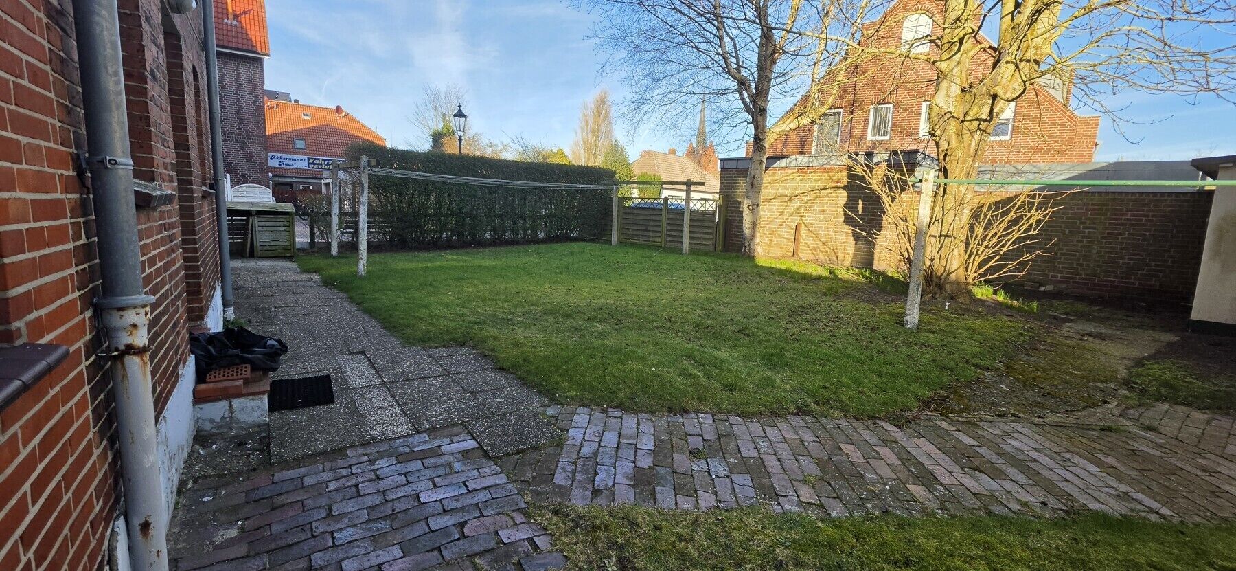 Un jardin avec une zone dallée, une clôture en bois et un bâtiment en briques.