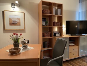 Ein modernes Wohnzimmer mit einem Holz-TV-Schrank, einem Flachbildfernseher, einem Bücherregal, einem Esstisch mit einer Tulpenvase und einem gerahmten Bild an der Wand.