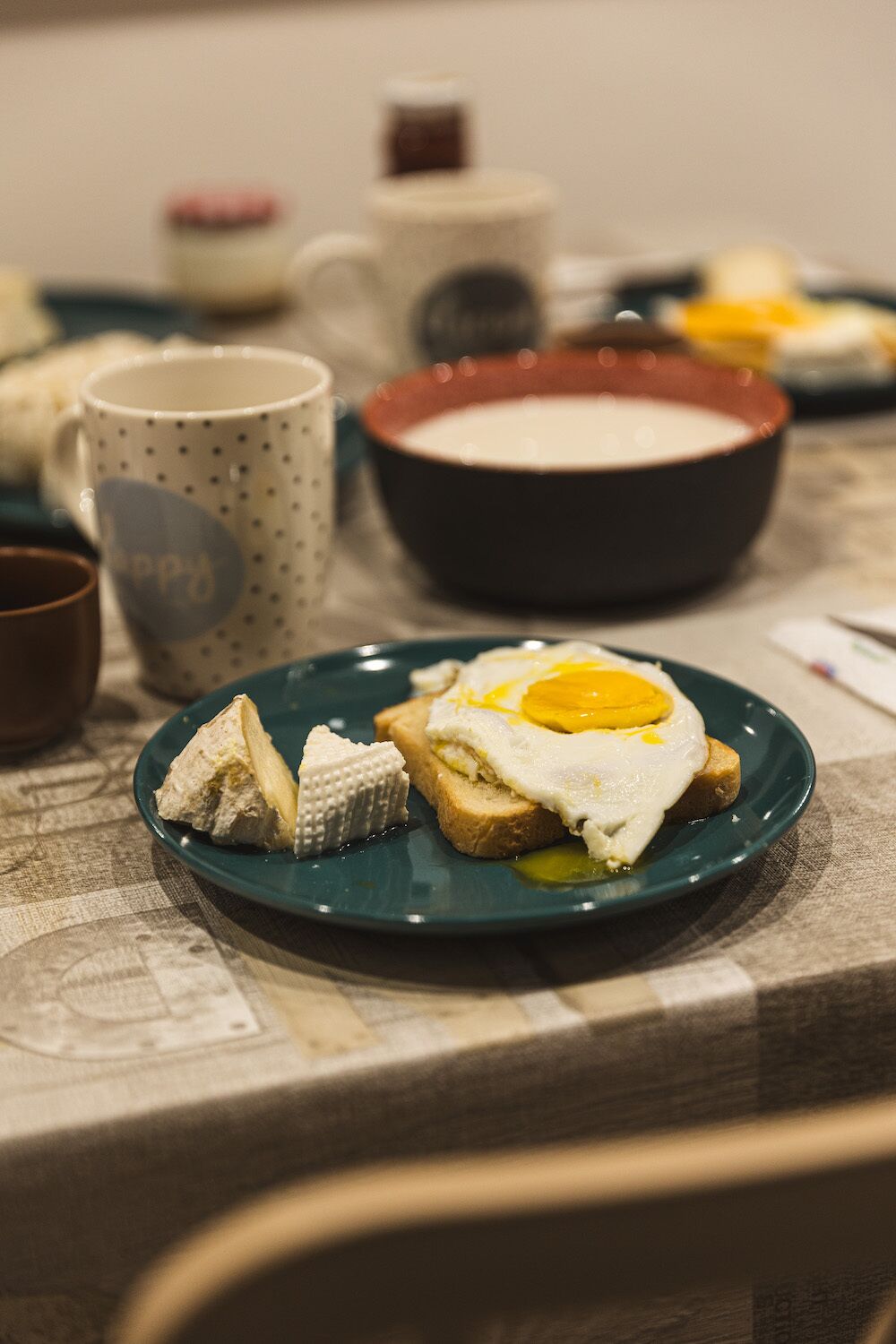 Une assiette avec un œuf au plat et du fromage sur une tranche de pain grillé, un bol de soupe et une tasse à pois sur une table.