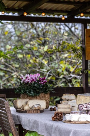 Una mesa con varios quesos y una cesta de flores, ubicada al aire libre bajo una estructura de madera.