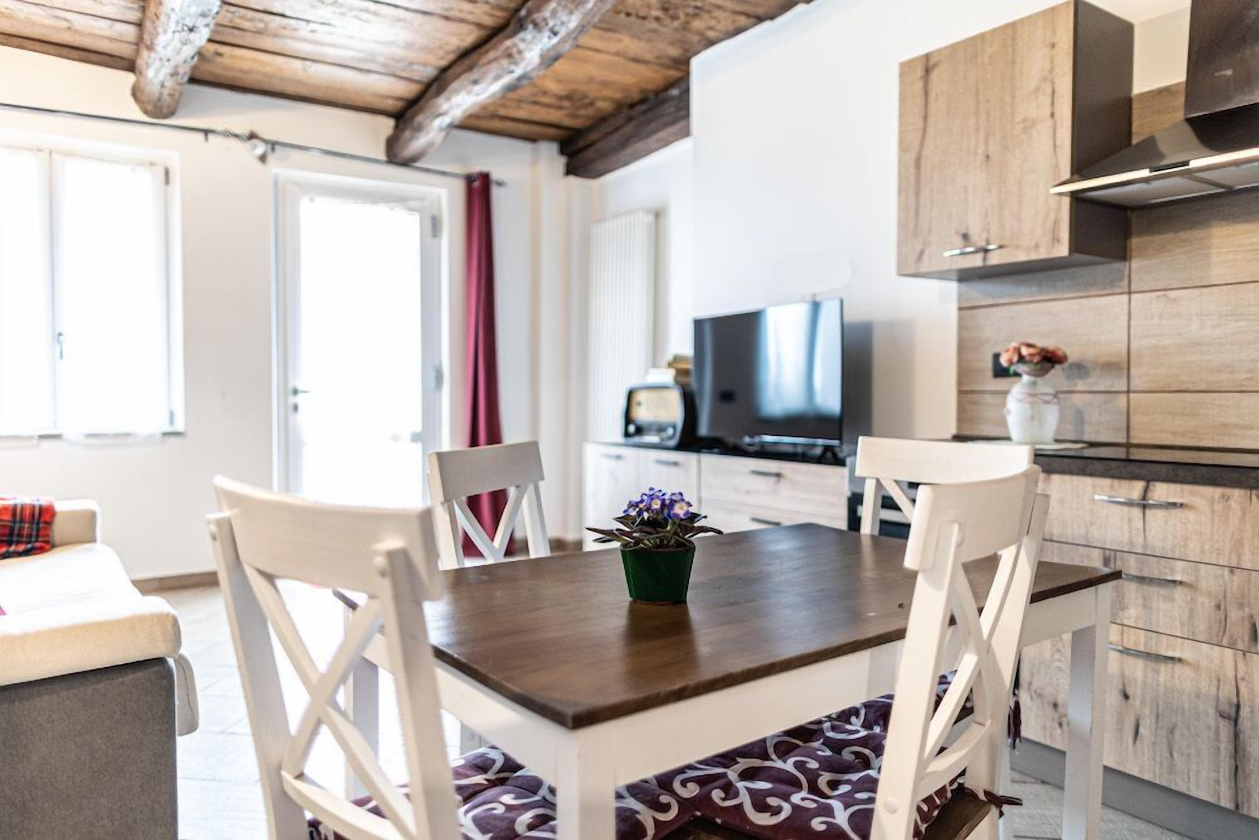 Un comedor con una mesa de madera y sillas blancas, un televisor al fondo y una cocina con armarios de madera.
