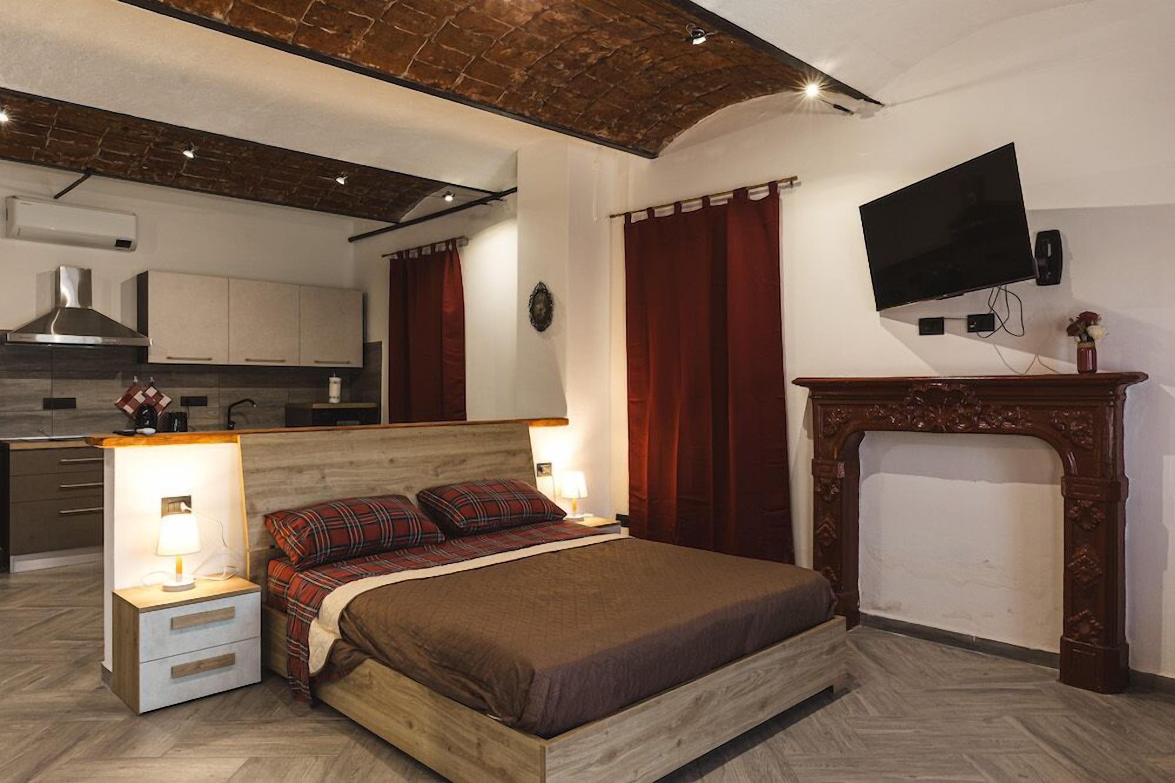 Una camera da letto con un letto di legno, un comodino con una lampada, un camino, una televisione e una cucina sullo sfondo.