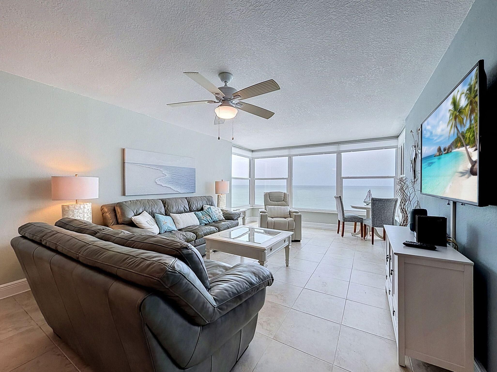 A spacious living room with a leather sofa, a coffee table, a ceiling fan, a TV, and a view of the ocean.