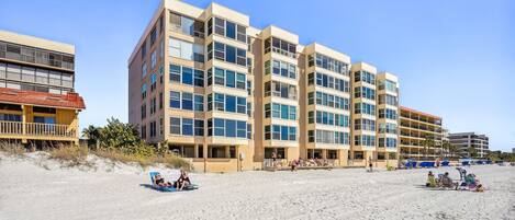 A beachfront hotel with multiple floors, balconies, and a sandy beach area with people and beach chairs.