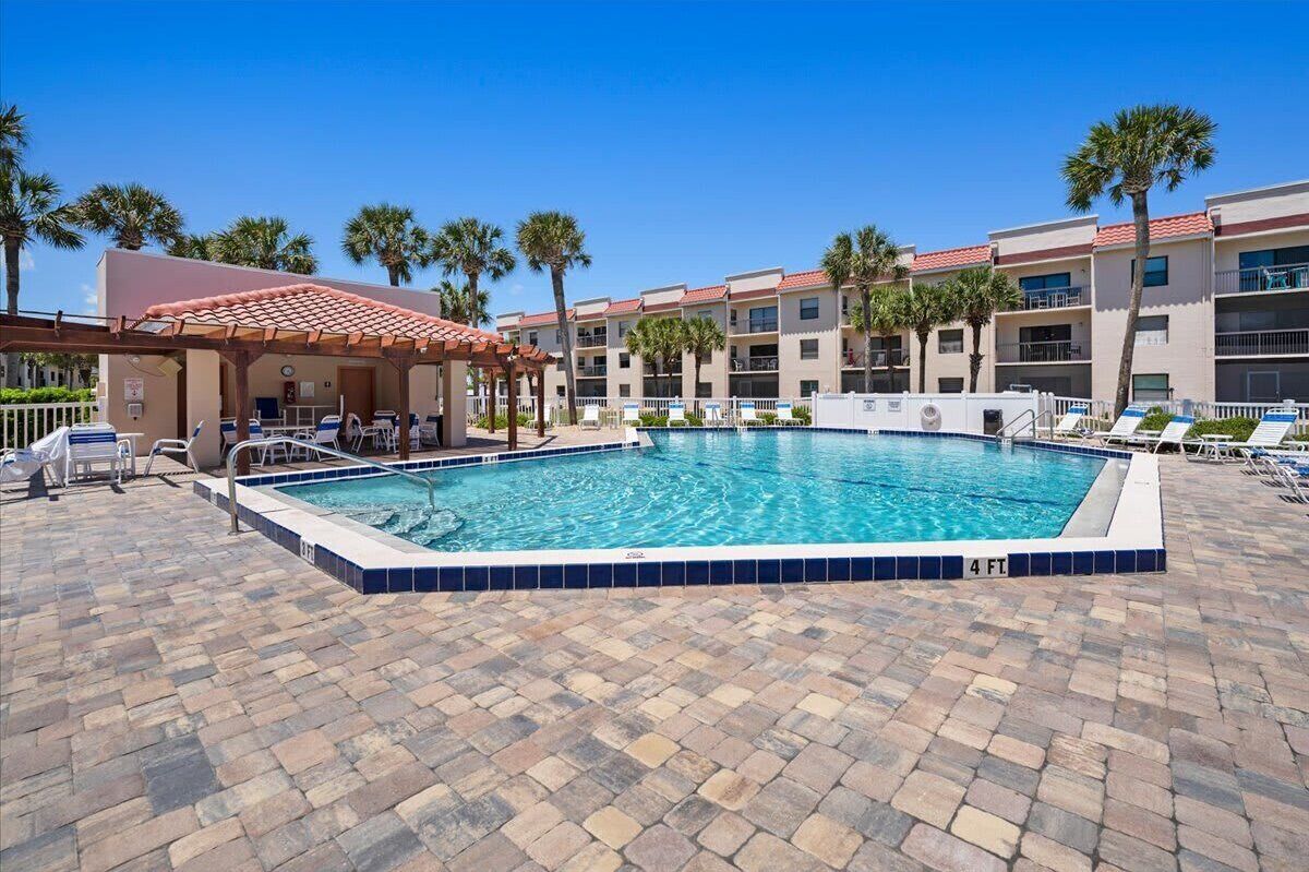 A swimming pool with a tiled border, a covered seating area, and a building with balconies in the background.