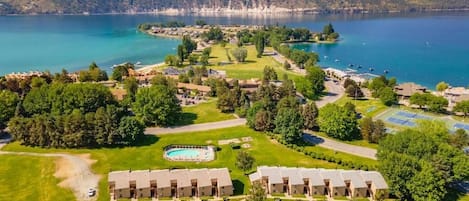 An aerial view of a resort with multiple buildings, a swimming pool, and a lake with mountains in the background.