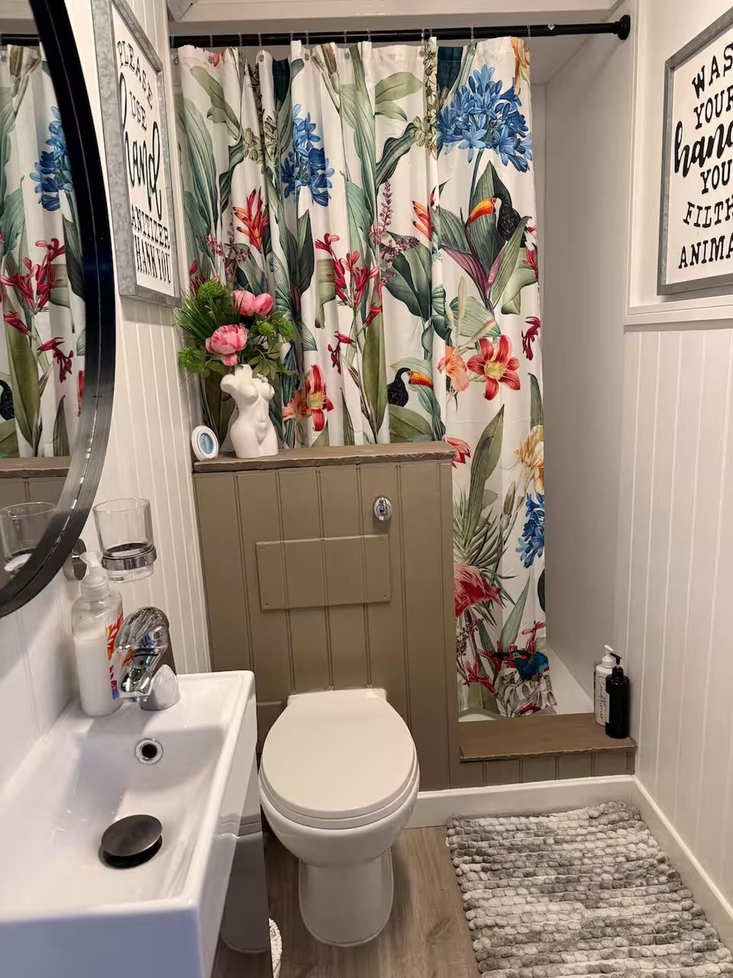 Une salle de bain comprenant des toilettes, un lavabo, un miroir, un rideau de douche à motifs floraux et un petit tapis.