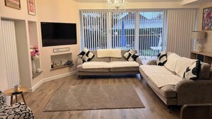 A living room with a sofa, a TV, a chandelier, and a large window with blinds. - Tranquil Solihull Retreat - 3 Bedroom House (Solihull)