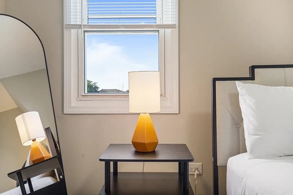 A hotel room with a bed, a bedside table with a lamp, a window with blinds, and a wall-mounted mirror.