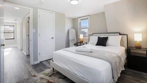 A modern bedroom with a large bed, bedside lamps, a window, and a door leading to another room. - Spacious 5 Bed 2 bath House (Melrose Park)