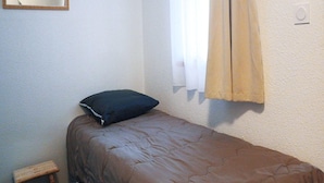 A small, single-bed room with a brown bedspread, a dark pillow, a small wooden stool, and a window with beige curtains. - Functional Studio, Heart of the High Pyrenees (Garin)