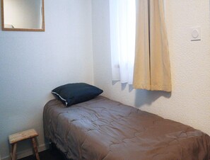 Ein kleines Zimmer mit einem Einzelbett, einer braunen Bettdecke, einem dunklen Kopfkissen, einem kleinen Holzschemel und einem Fenster mit beigen Vorhängen.