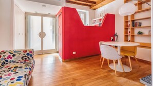 Une pièce avec un plancher en bois, un mur d’accent rouge, une table ronde blanche, une chaise blanche, un canapé coloré et une bibliothèque.