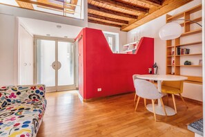 Ein Zimmer mit Holzboden, einer roten Akzentwand, einem weißen runden Tisch, einem weißen Stuhl, einem bunten Sofa und einem Bücherregal.