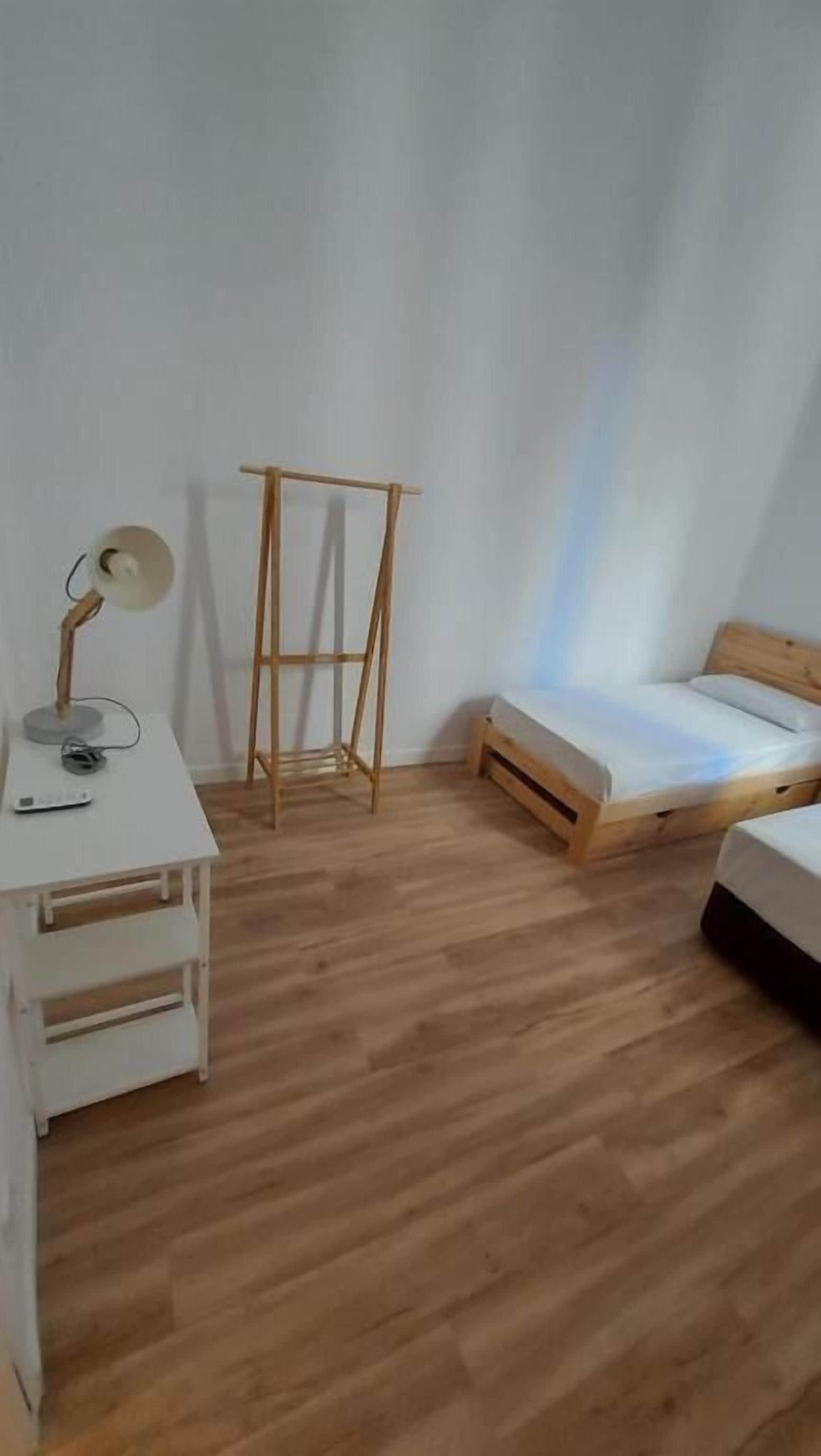 A room with a wooden floor, a white desk with a lamp, a bed with a wooden frame, and a standing fan.