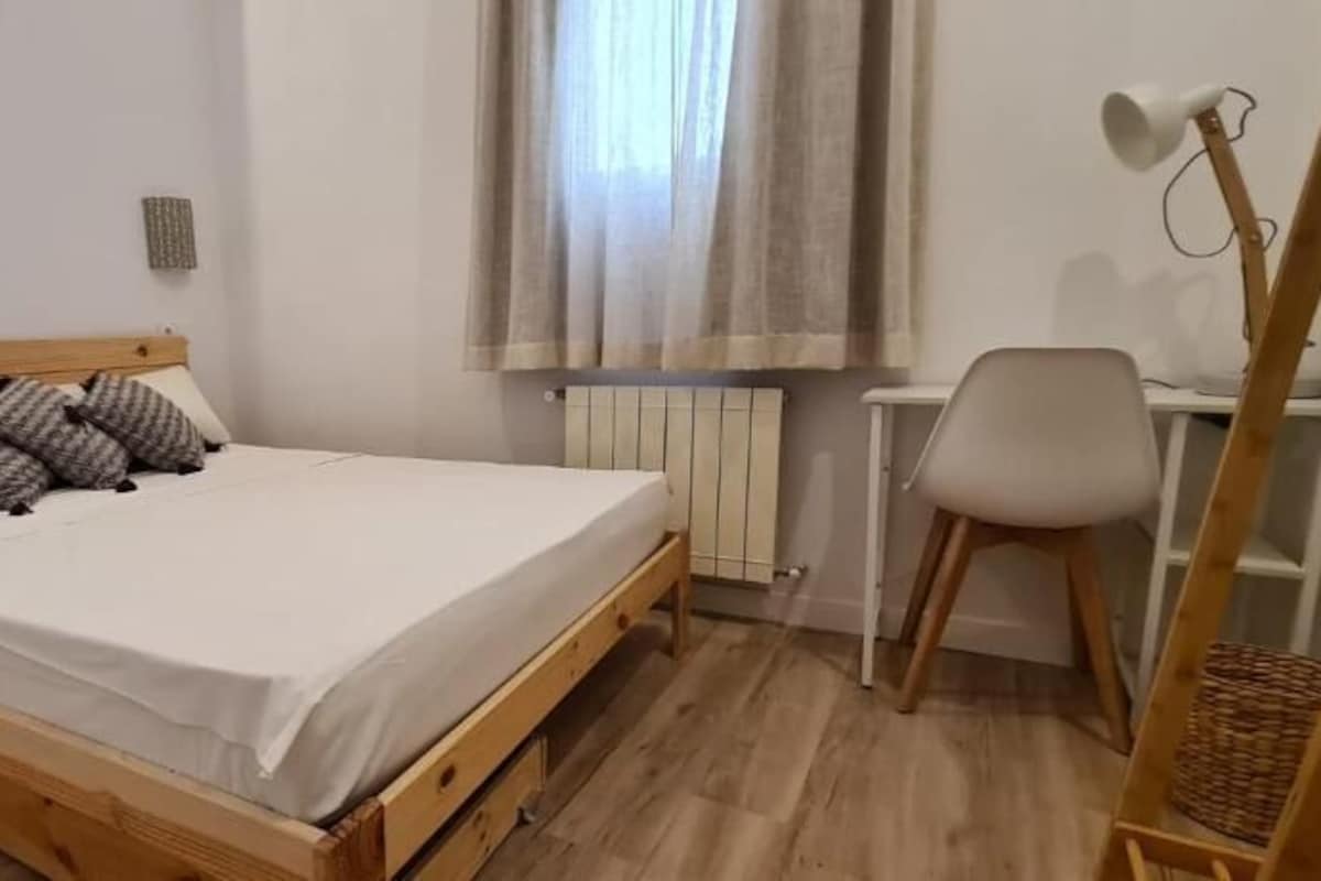 A bedroom with a wooden bed, a white chair, a desk, and a radiator.
