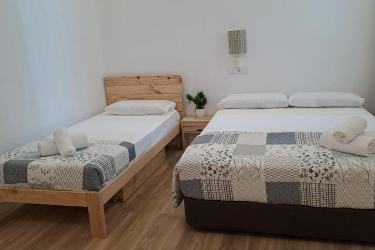 A room with two single beds and a larger bed, wooden headboards, a small potted plant, and a wall-mounted light fixture.
