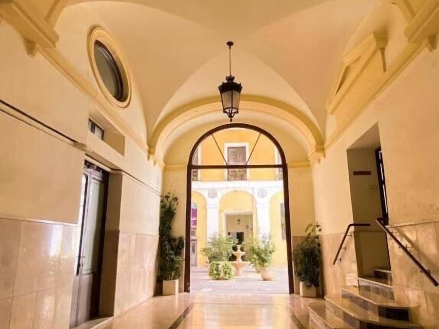 Un corridoio con archi, un lampadario al centro e una scala che conduce a un piano superiore.