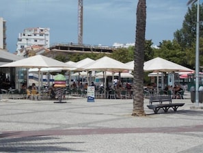 Een buitenzitplaats met parasols, tafels en stoelen, omgeven door palmbomen en gebouwen.
