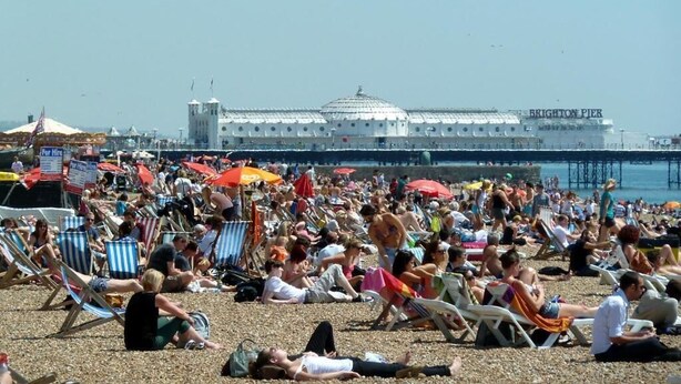A crowded beach with people relaxing on sun loungers and umbrellas, a pier in the background, and a large building with a dome roof. - Number 75 (Brighton)