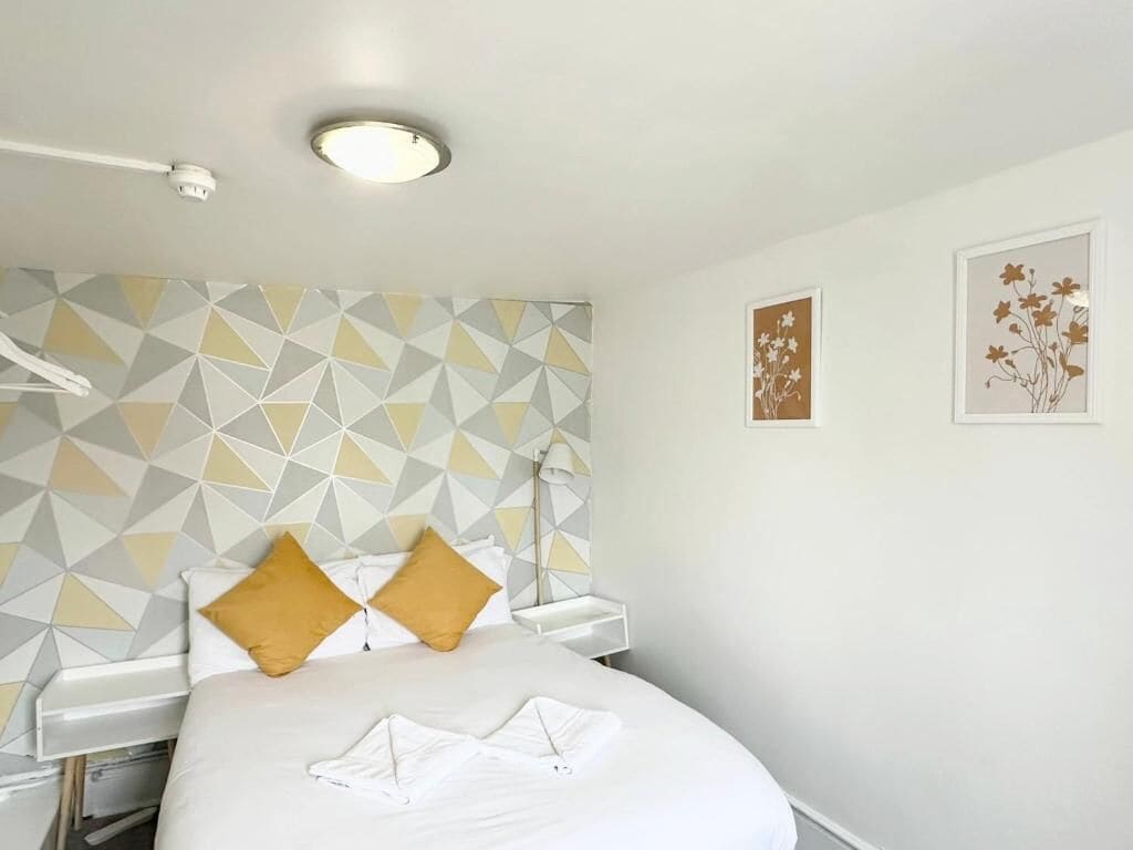 A hotel room with a bed, a geometric-patterned wall, a ceiling light, and two framed artworks.