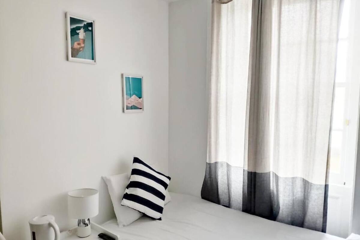 Un lit simple avec un oreiller à rayures blanches et grises, une table de chevet avec une bouilloire et une lampe, et deux tableaux encadrés accrochés au mur.