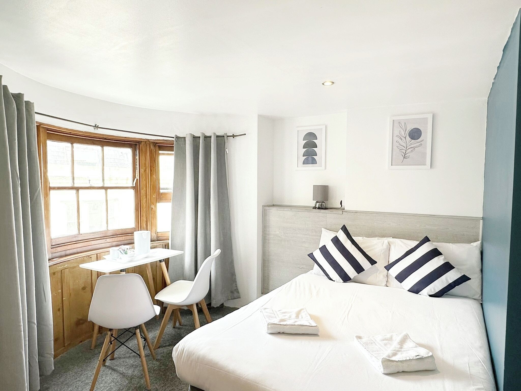 A hotel room with a bed, a desk, two chairs, a window with curtains, and wall art.