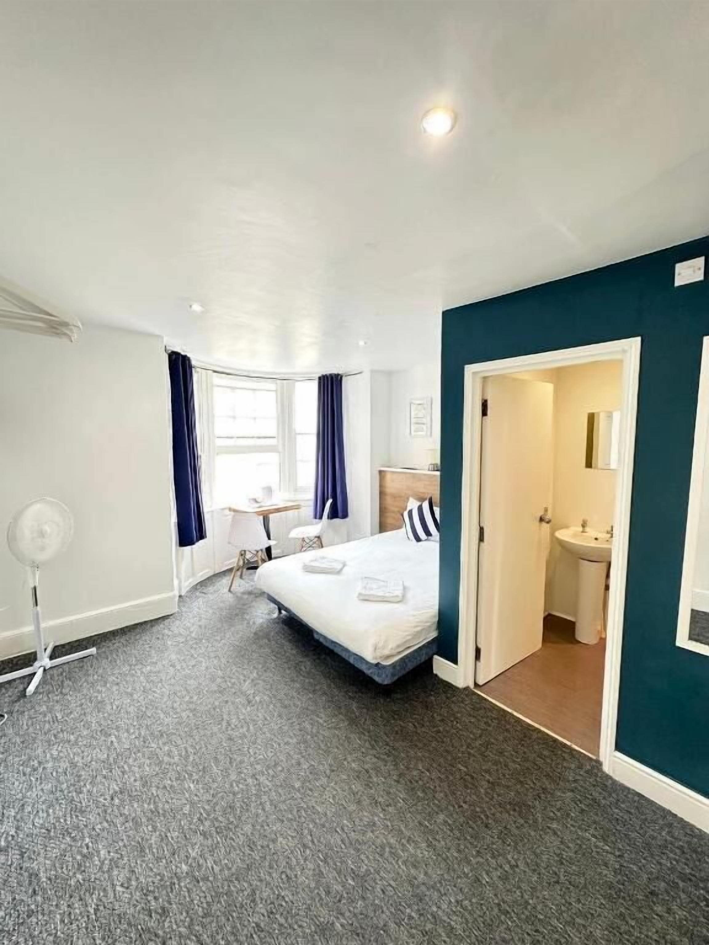 A hotel room with a bed, a desk, a chair, a fan, and a bathroom.