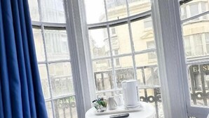 A corner room with a bay window, a white table with a remote control, two white chairs, and a blue curtain. - Number 75 (Brighton)