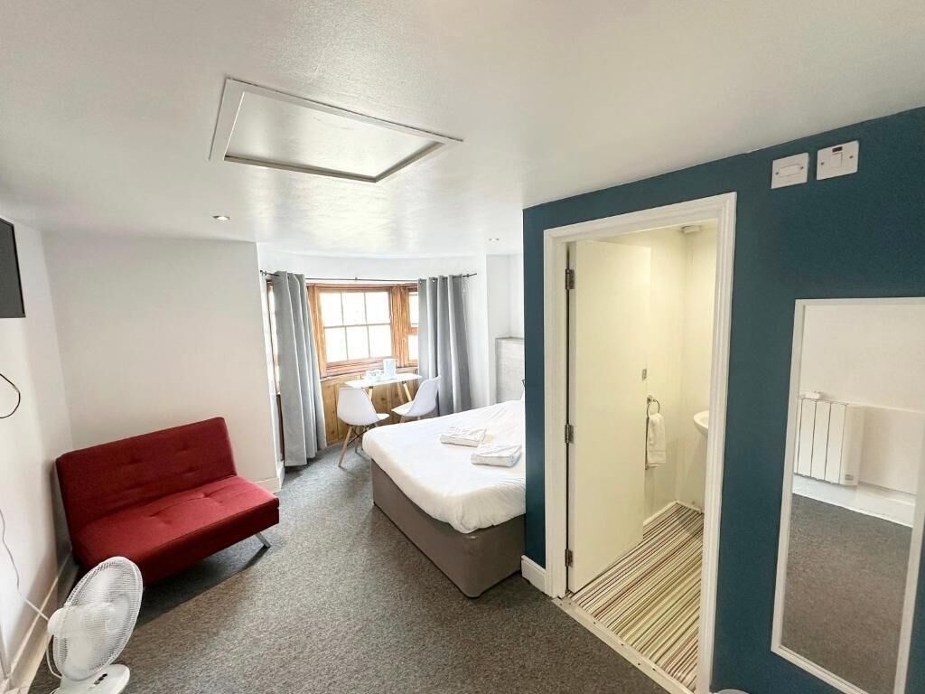 Habitación de hotel con cama, sofá rojo, ventilador, ducha y vistas al exterior.