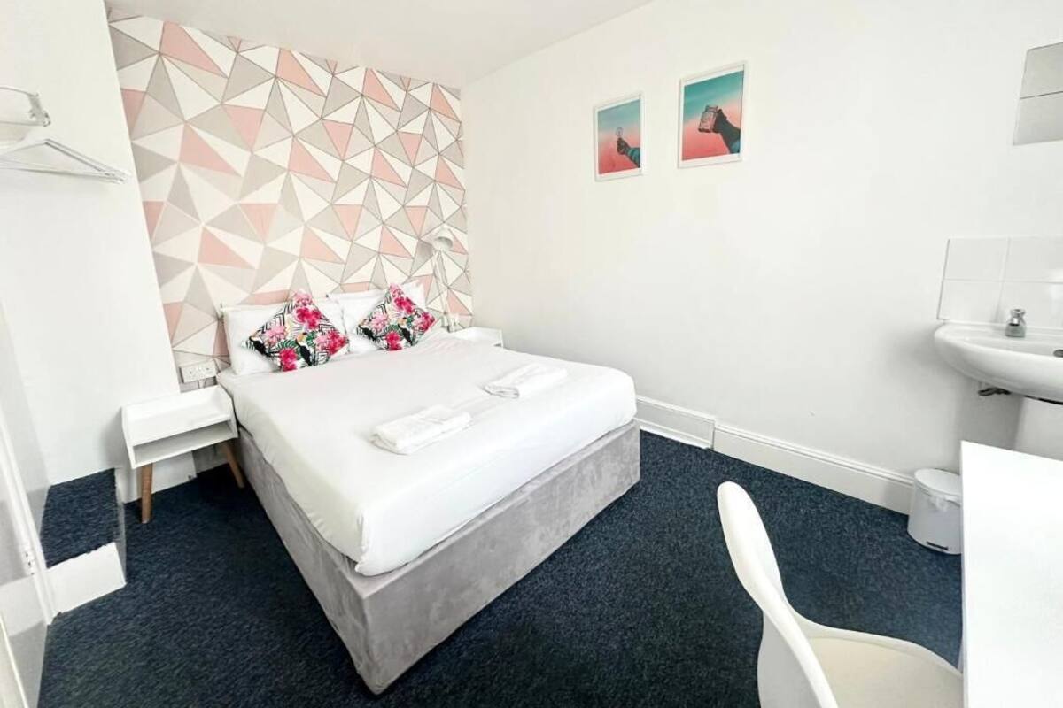 Une chambre d’hôtel comprenant un lit, une table de chevet, une chaise, un mur orné de motifs géométriques, ainsi qu’une salle de bain avec un lavabo et un miroir.