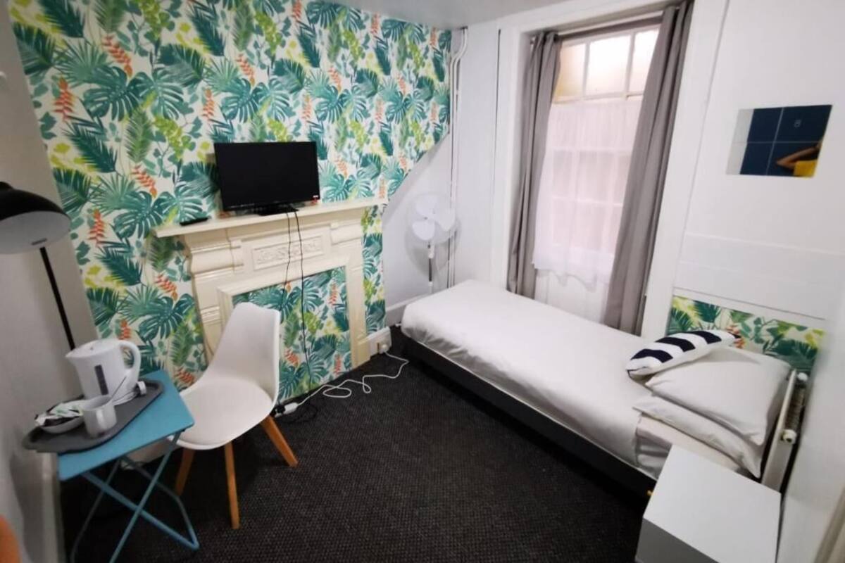 Une petite chambre d’hôtel avec un lit, un bureau avec une télévision, une chaise, une fenêtre avec des rideaux et un papier peint aux motifs tropicaux.