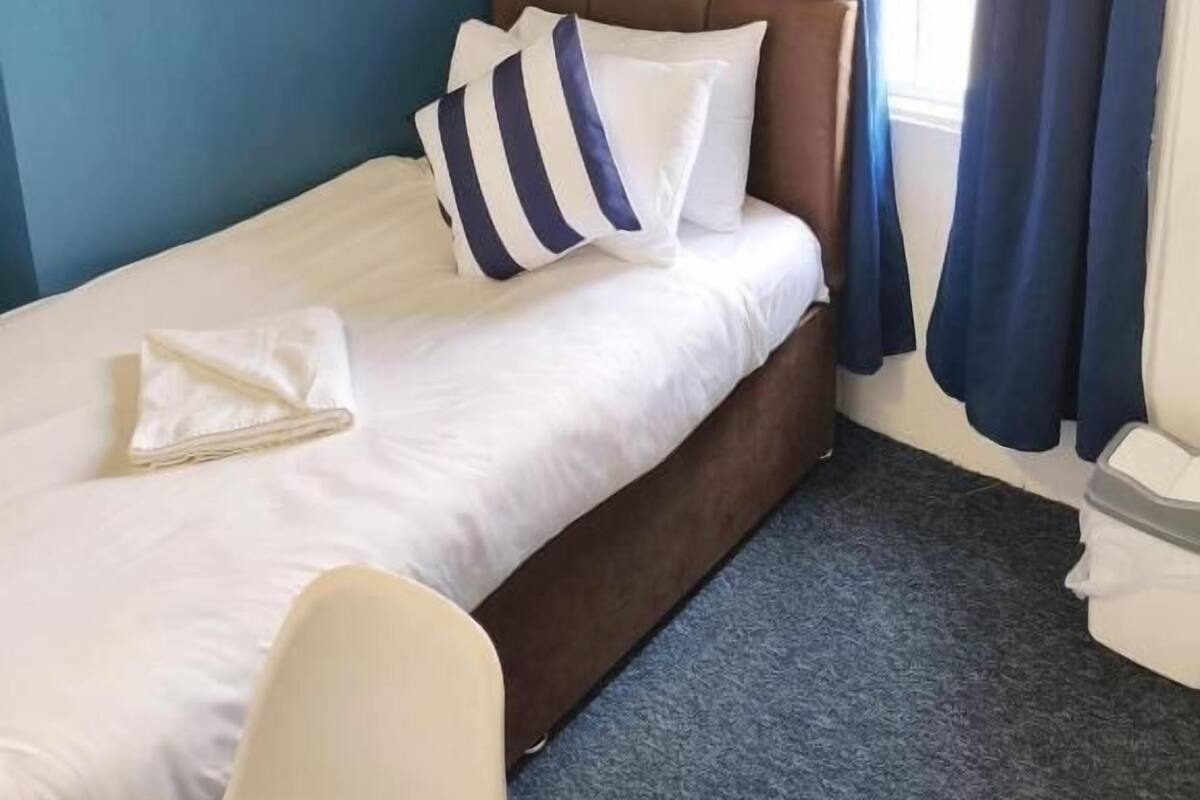 Un lit simple avec une tête de lit, un oreiller, une serviette pliée et une décoration en forme de roue de navire accrochée au mur.