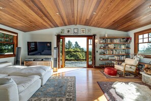 A spacious living room with a wooden ceiling, a large TV, a sofa, a red ottoman, and a bookshelf filled with books. - Horizon House | Hot Tub, Views, Gardens + Golf (Sebastopol)