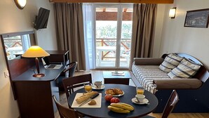 Ein Zimmer mit Esstisch, gedecktem für ein Frühstück, einem Schreibtisch mit Laptop, einer Couch mit Kissen und einem Balkon mit Vorhängen.