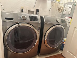 Ein Waschraum mit zwei Samsung Waschmaschinen, einem Durchlauferhitzer und einem an der Wand befestigten Regal mit Reinigungsmitteln.