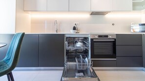 A modern kitchen with a dishwasher, oven, and a dining area with a chair. - Rooftop Glasshouse Retreat by LovelyStay (Porto)