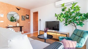 En moderne stue med sofa, fjernsyn, en plante og en vægmonteret aircondition.