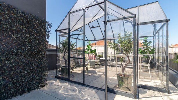 Et drivhus med glasvægge, en fliseterrasse og plantede planter indeni.