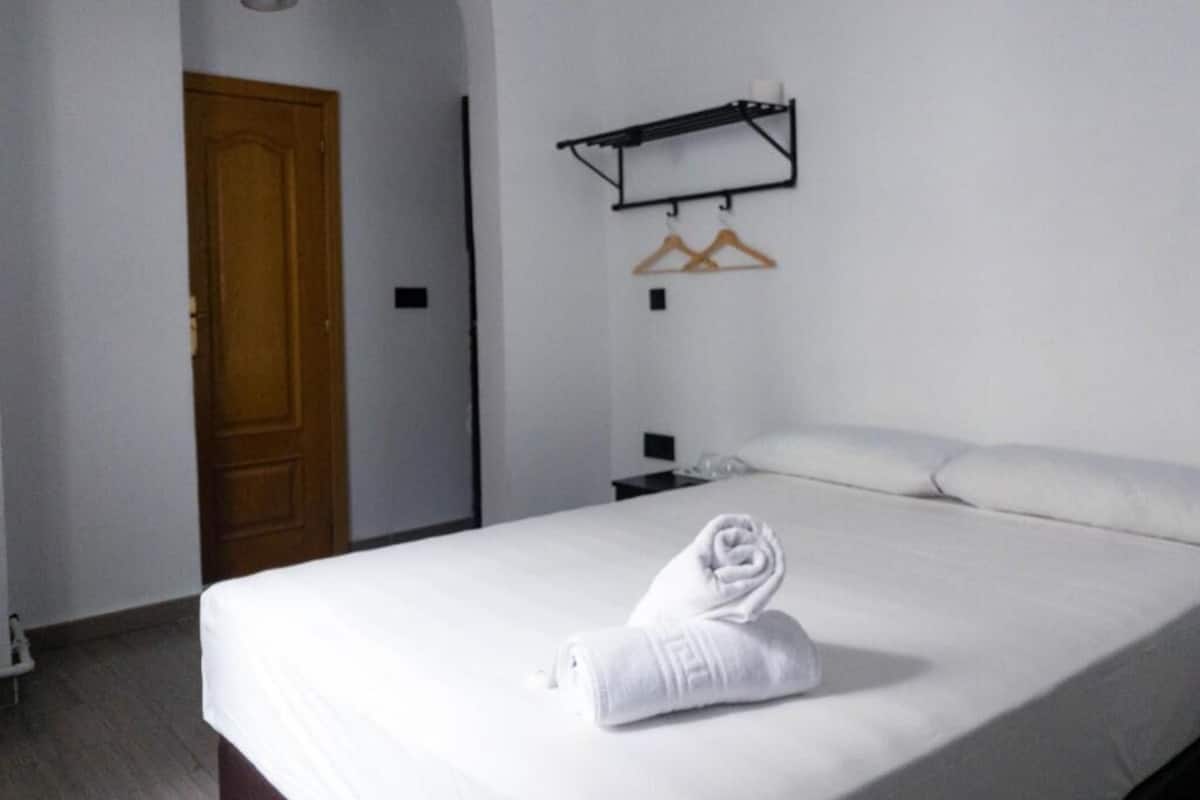 A hotel room with a bed, a wooden door, a hanging light fixture, a black shelf with hangers, and a white towel on the bed.