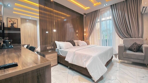 A modern hotel room with a wooden desk, a large bed, a grey armchair, and a large window with curtains. - Wealth's Khort Apartment (Keffi)
