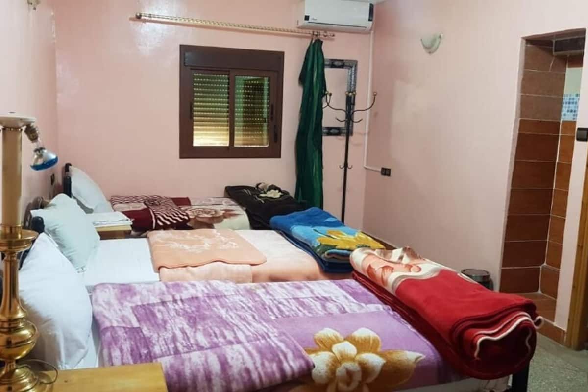 Una habitación con cama, mesita de noche, lámpara, ventana con persianas y cortina, y pared con azulejos.
