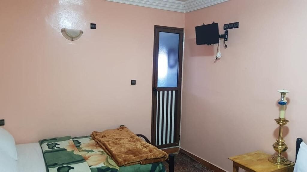 A room with a bed, a small table with a lamp, a wall-mounted TV, and a door with a window.