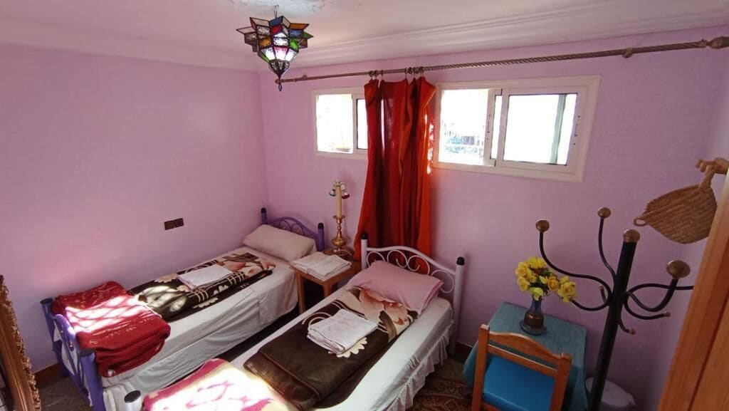 Una habitación pequeña con dos camas, una mesa con un florero, una silla y una lámpara colgante.