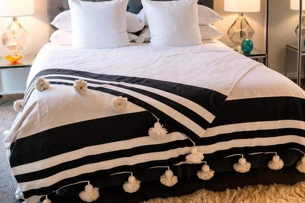 Una cama con cabecera capitonada, ropa de cama blanca y almohadones a rayas con pompones.