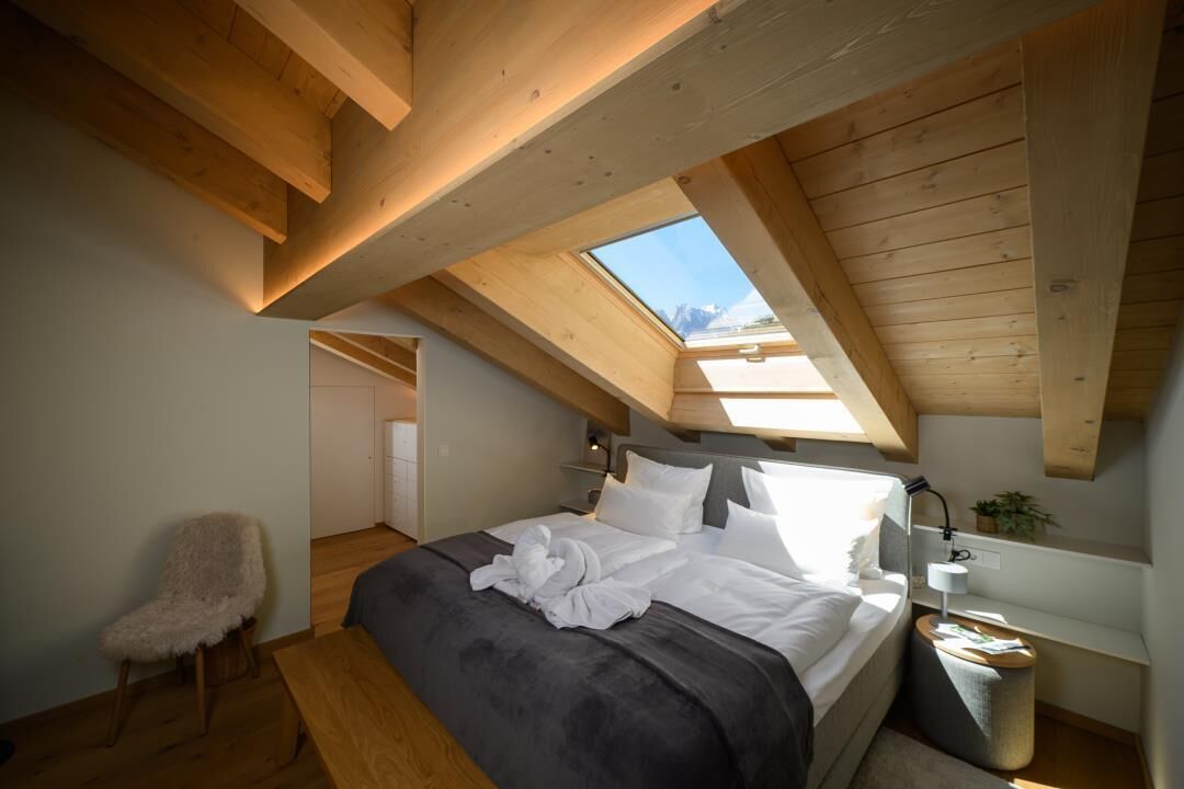 Ein Schlafzimmer mit Oberlicht, Holzbalken, einem Bett, einem Nachttisch und einem Sessel.