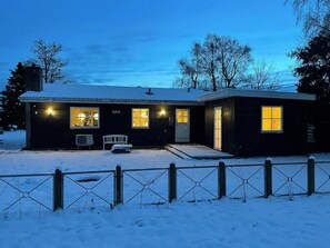 Een huis met twee verdiepingen, een besneeuwde voortuin en een omheinde tuin, met een bankje.