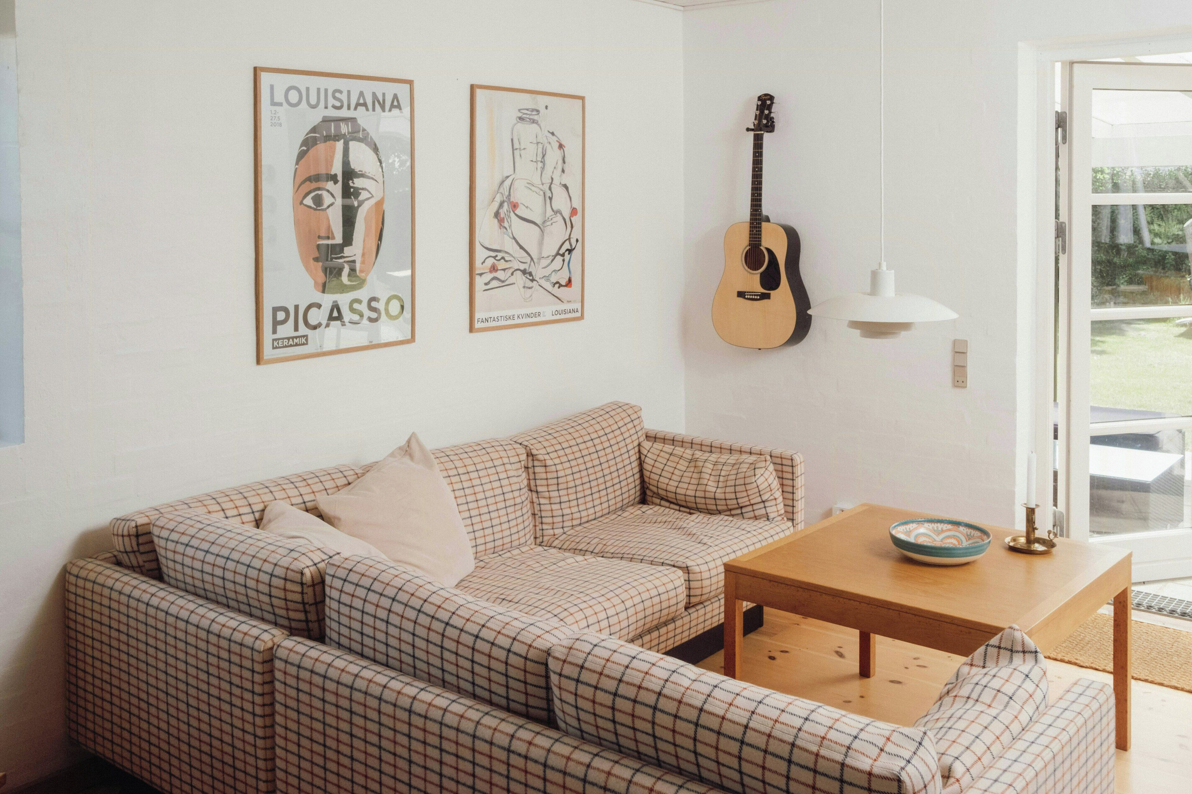 A living room with a checkered sofa, a guitar, a table with a bowl, and two framed artworks on the wall.